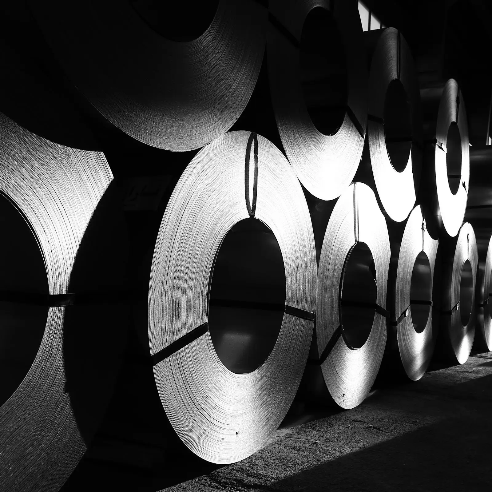 A long row of steel coils standing on edge in a dim warehouse, light catching their wound edges.