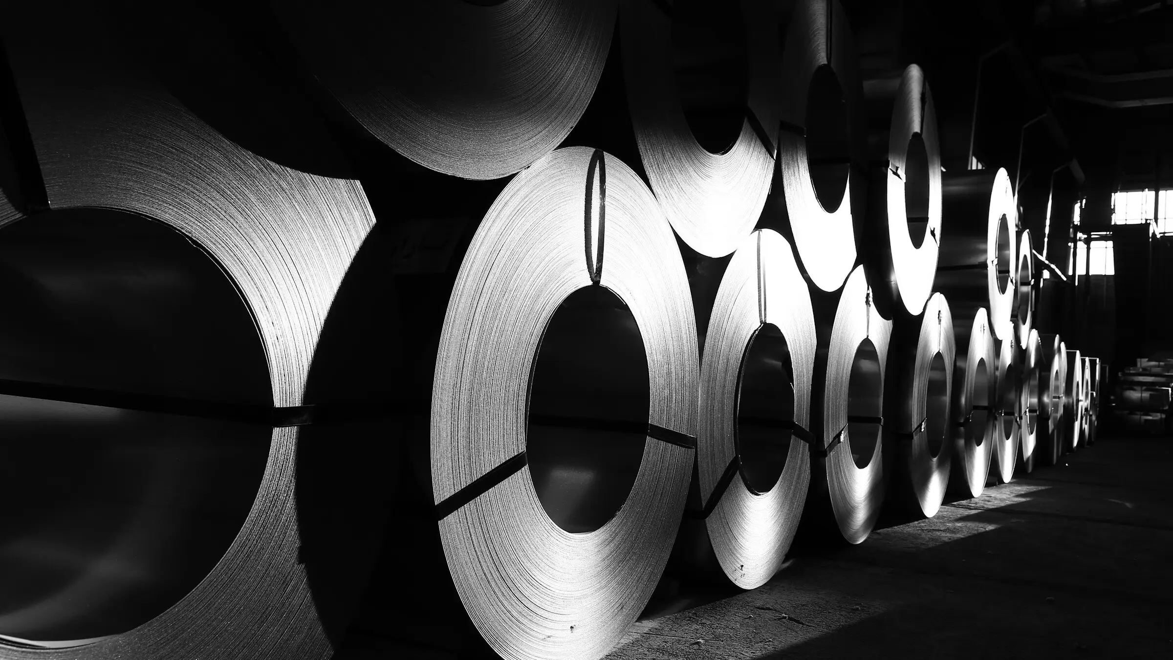 A long row of steel coils standing on edge in a dim warehouse, light catching their wound edges.