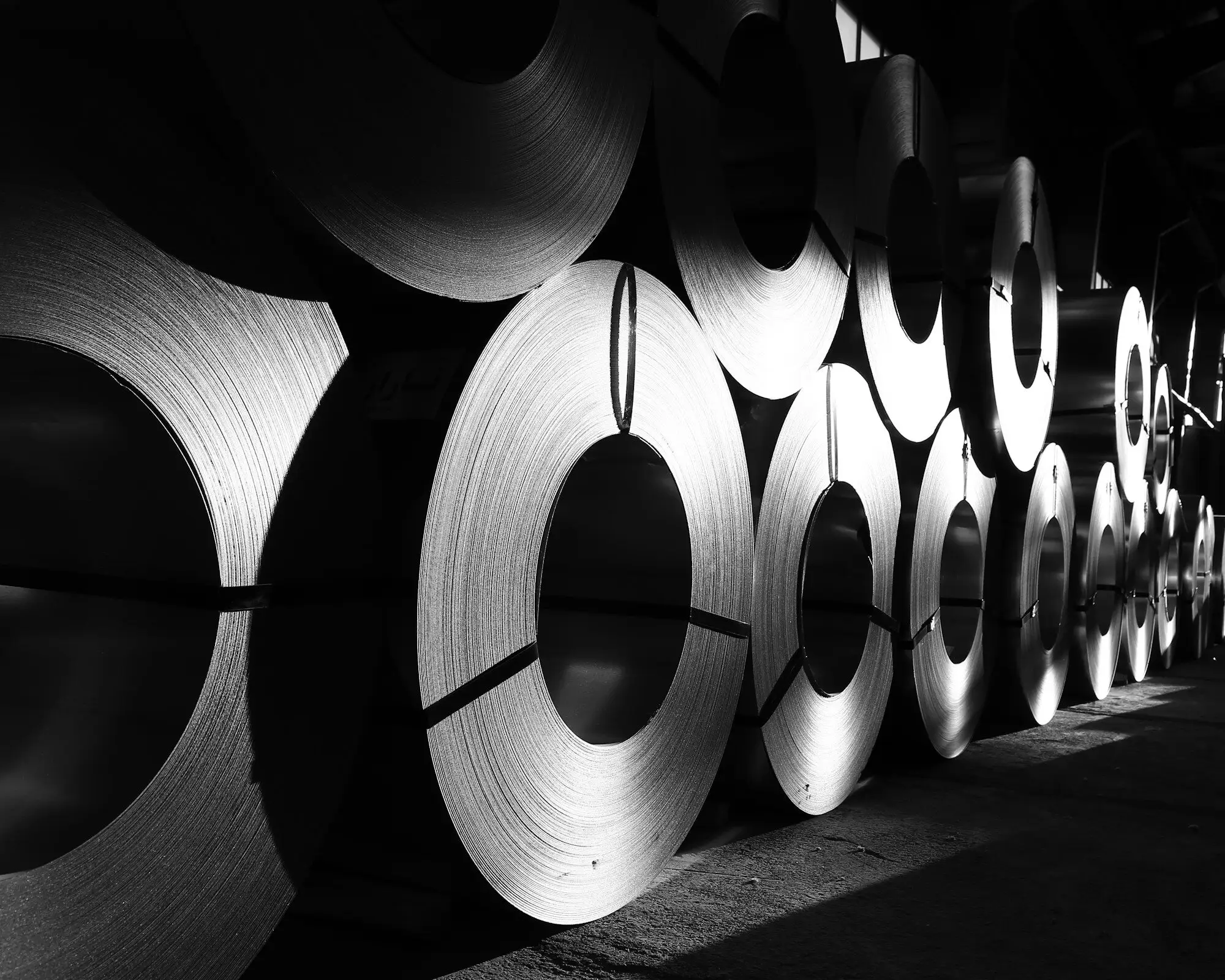 A long row of steel coils standing on edge in a dim warehouse, light catching their wound edges.