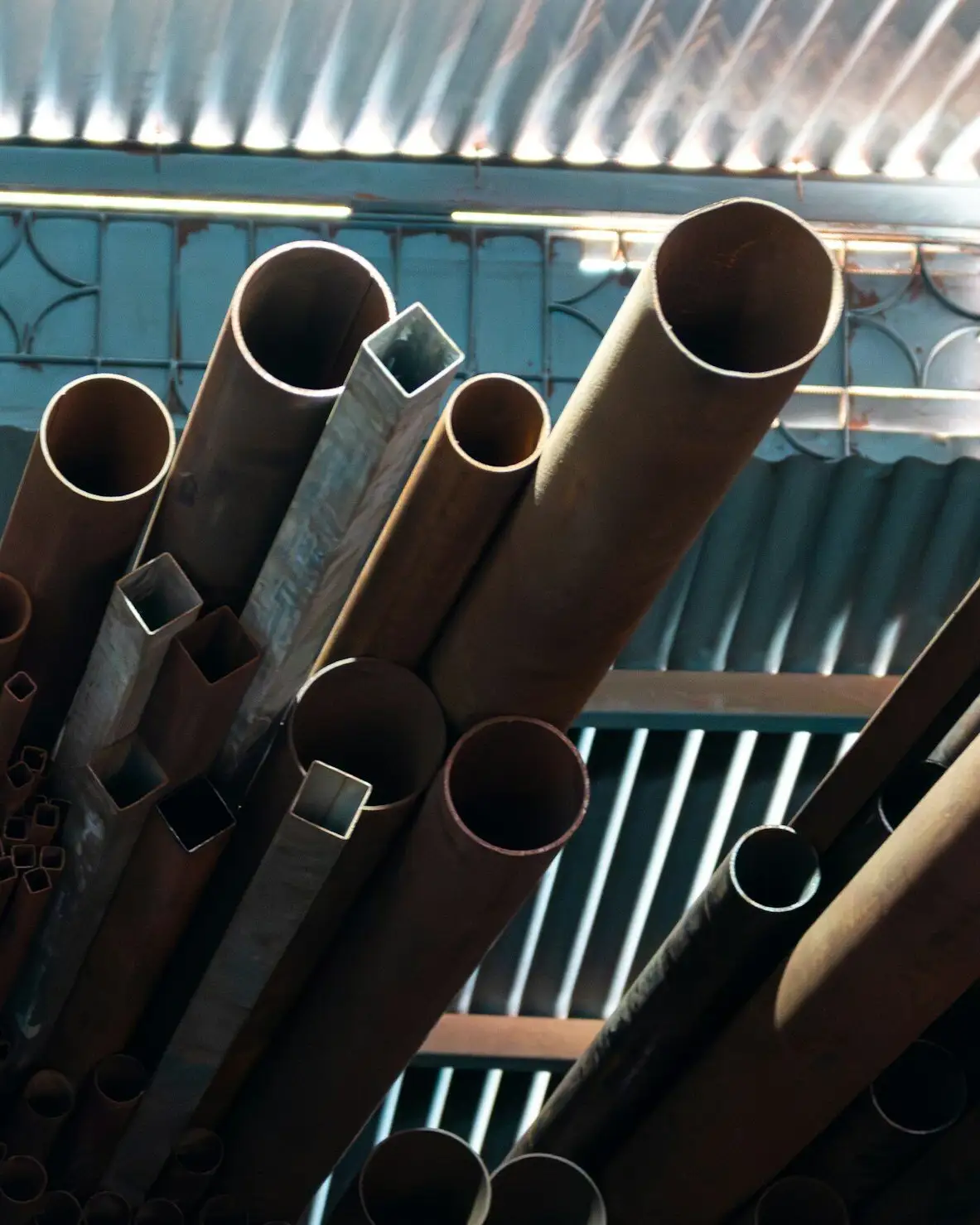 Round and square steel pipes standing in a stockist’s rack under a corrugated roof.