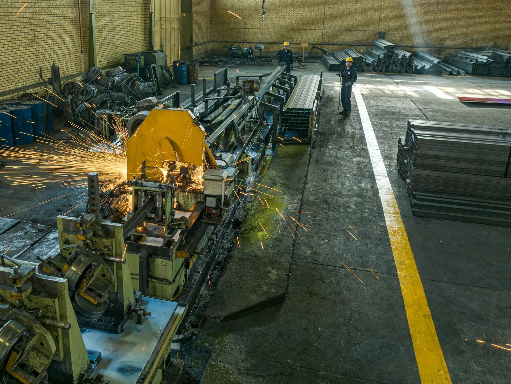 A tube mill line running in a brick-walled shed, sparks flying from the cut-off saw while two operators in hard hats watch the line.