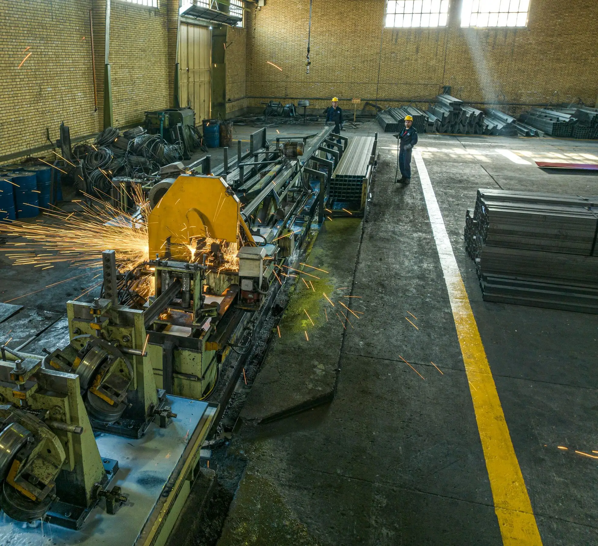 A tube mill line running in a brick-walled shed, sparks flying from the cut-off saw while two operators in hard hats watch the line.