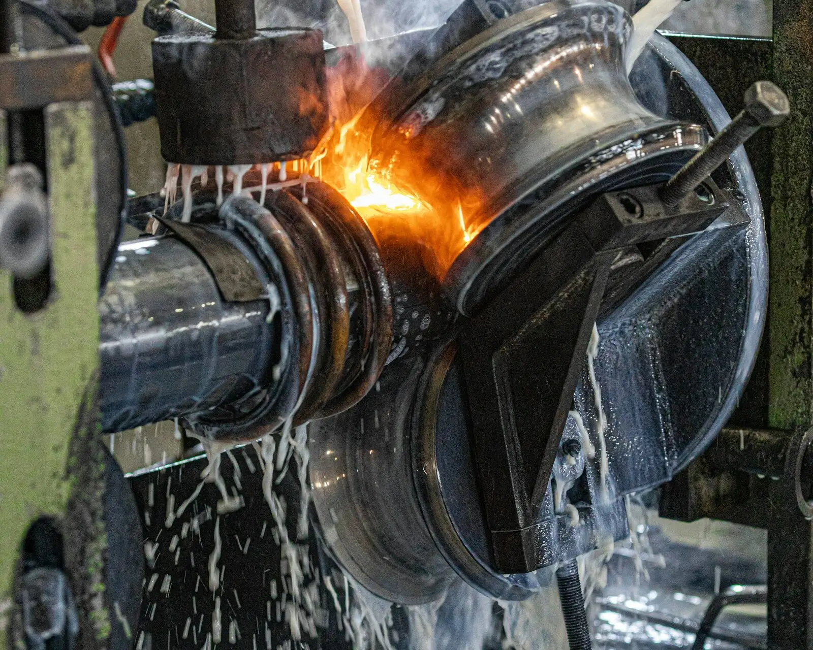 Close-up of a tube mill weld station: the strip glows orange at the seam between steel squeeze rolls while coolant pours over them.