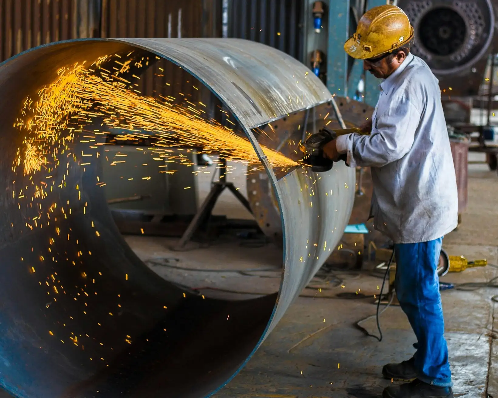A worker in a hard hat and safety glasses grinding a steel shell, sparks spraying across the workshop.