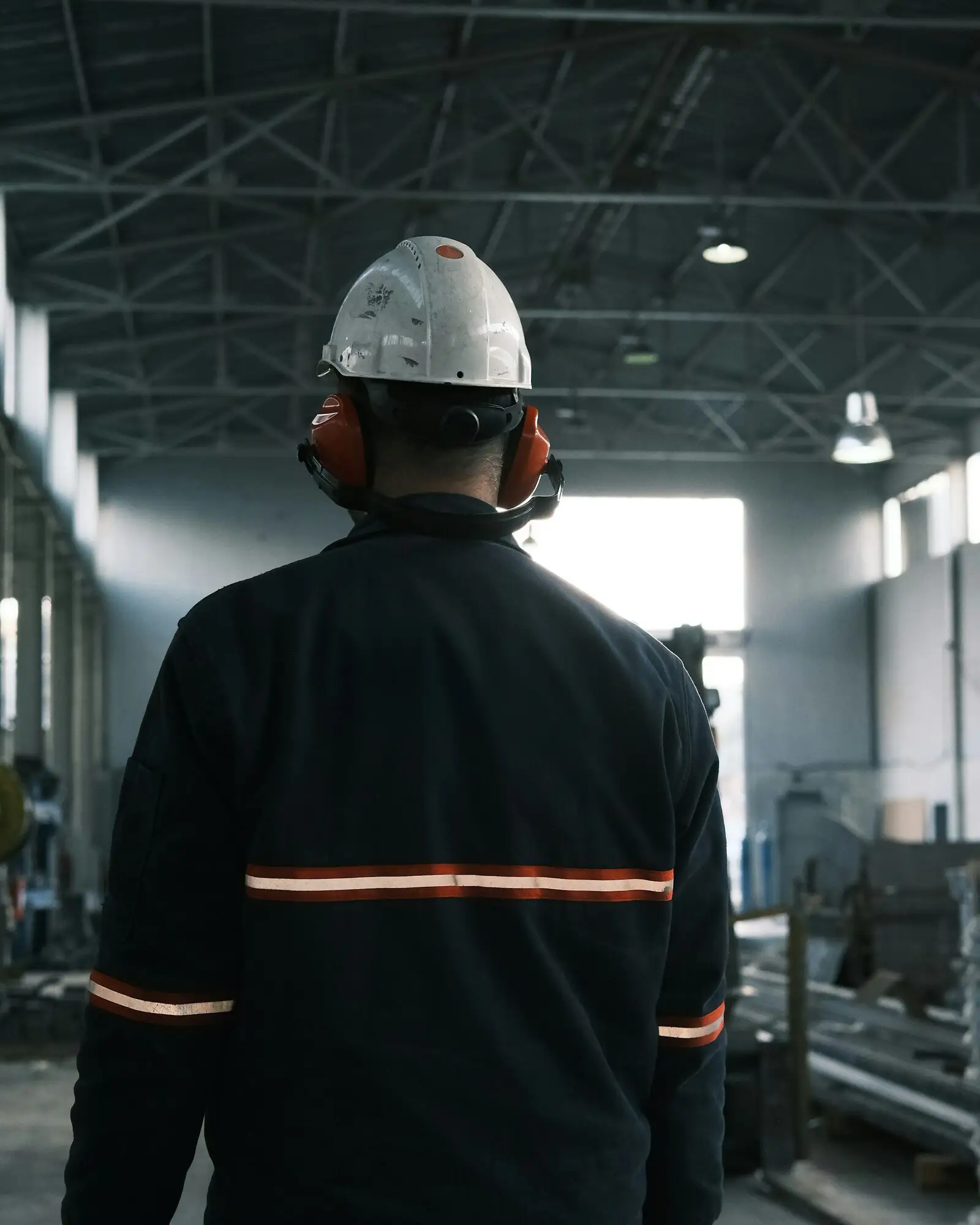 A mill worker seen from behind in a hard hat, ear defenders and reflective workwear, walking down a factory bay.